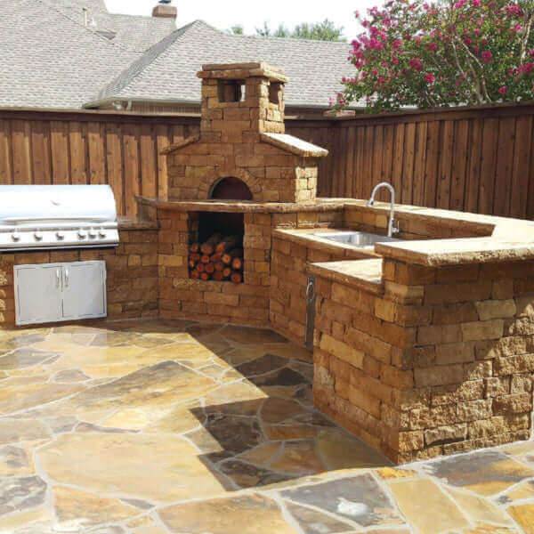 Outdoor masonry pizza oven and grill in a backyard setting with stone patio and wooden fence.